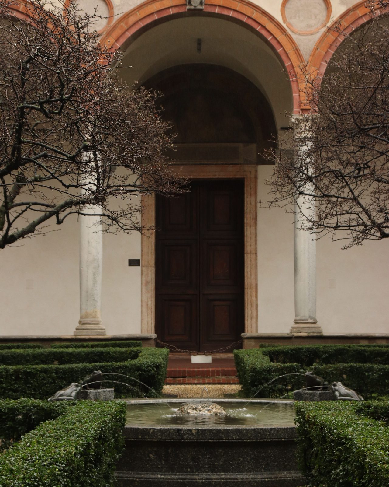 Elegant courtyard featuring a stone fountain, trimmed hedges, and an arched entrance with intricate columns.