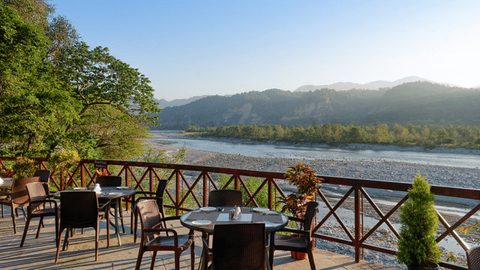 Outdoor dining deck with tables overlooking the Kosi River and hills at Infinity Resorts Corbett